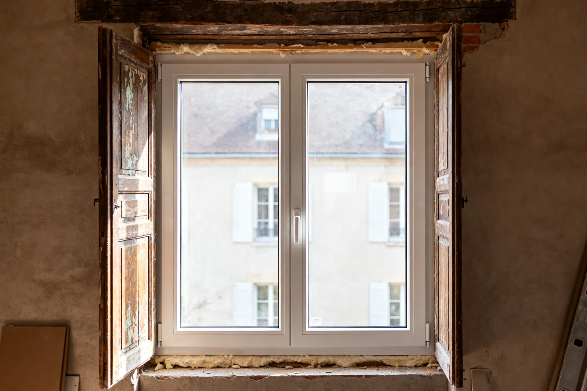 Vue d'une fenêtre en rénovation avec volets en bois et cadre moderne PVC.