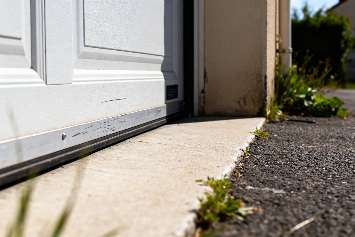 Vue rapprochée d'un seuil alu porte de garage sur sol béton avec herbes.