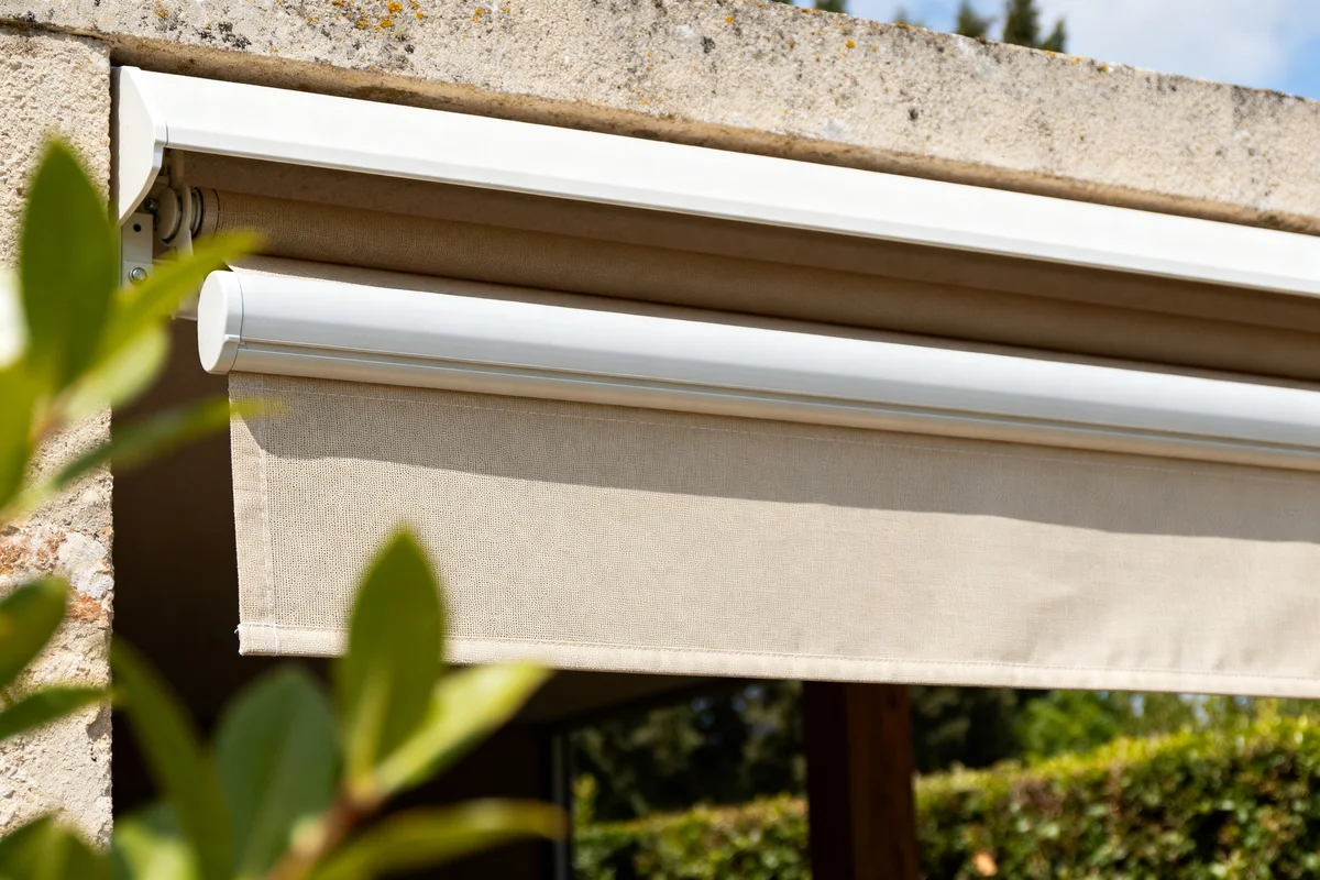 Store banne tissus beige installé sur une terrasse ensoleillée devant des arbustes verts